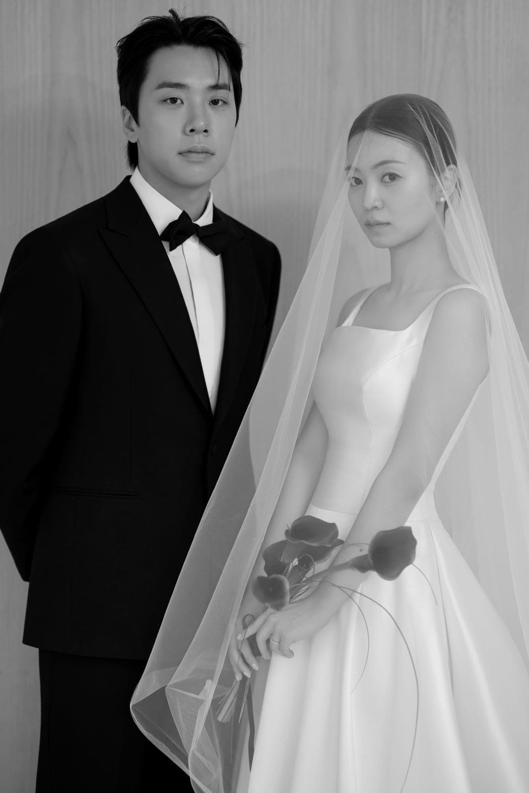 흑백 사진. 턱시도를 입은 신랑과 베일을 쓴 신부가 나란히 서서 정면을 바라보는 모습