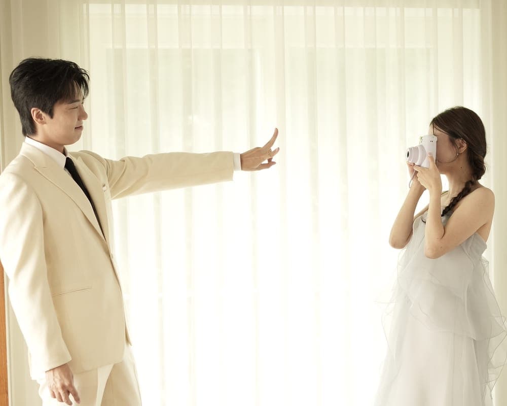 신부가 즉석카메라로 신랑을 찍고, 신랑이 손을 뻗어 가리는 모습